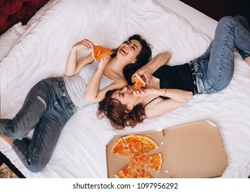 The stylish sisters eating slices of pizza and lying on the bed