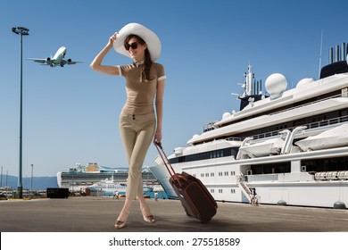Stylish slim woman wearing sunglasses and white hat standing in seaport near docked cruise ship while smiling and holding her luggage on a summer sunny day. Travel, vacation, adventure concept.