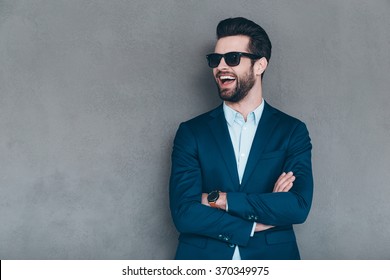 Stylish Smile. Cheerful Young Handsome Man In Sunglasses Keeping Arms Crossed And Looking Away With Smile While Standing Against Grey Background