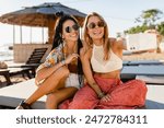 stylish smiling beautiful women relaxing having fun on beach in summer fashion trend outfit, colorful apparel boho style, tropical vacation wearing sunglasses, using smartphone taking selfie picture