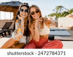 stylish smiling beautiful women relaxing having fun on beach in summer fashion trend outfit, colorful apparel boho style, tropical vacation wearing sunglasses, using smartphone taking selfie picture