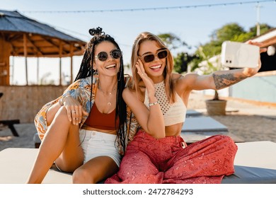 stylish smiling beautiful women relaxing having fun on beach in summer fashion trend outfit, colorful apparel boho style, tropical vacation wearing sunglasses, using smartphone taking selfie picture - Powered by Shutterstock