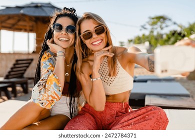 stylish smiling beautiful women relaxing having fun on beach in summer fashion trend outfit, colorful apparel boho style, tropical vacation wearing sunglasses, using smartphone taking selfie picture