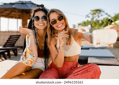 stylish smiling beautiful women relaxing having fun on beach in summer fashion trend outfit, colorful apparel boho style, tropical vacation wearing sunglasses, using smartphone taking selfie picture - Powered by Shutterstock