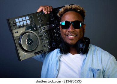Stylish smiling black man wears black glasses and denim shirt. Popular DJ with an earring in his ear and headphones carries DJ console on his shoulder to concert to entertain audience with music track