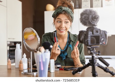 Stylish successful vlogger sitting at dressing room with cosmetic products, mirror and professional camera fixed on tripod, comparing two lipsticks in her hands, advertising famous brand via her vlog - Powered by Shutterstock