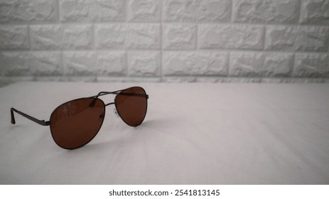 Stylish sunglasses with brown lenses and a sleek metal frame, placed on a white surface with a textured brick wall background. Perfect for fashion and accessory themes. - Powered by Shutterstock