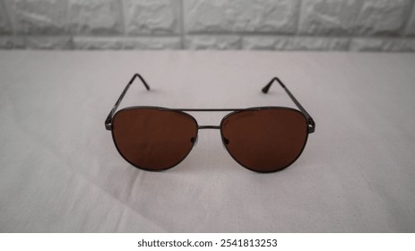 Stylish sunglasses with brown lenses and a sleek metal frame, placed on a white surface with a textured brick wall background. Perfect for fashion and accessory themes. - Powered by Shutterstock