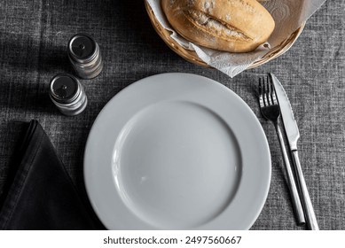 Stylish table beverage serving with white plate, black napkin, knife, fork, salt and pepper shekers, bread in busket on grey table clothe background