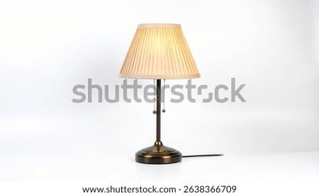 A stylish table lamp features a pleated beige fabric shade atop a bronze metal base. A simple design with subtle elegance. The lamp is illuminated and plugged in.