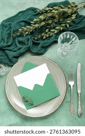 Stylish table setting, envelope with blank card and plant branches on grunge green background