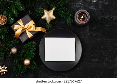 Stylish table setting, gift box, fir branches, star and balls, black plate with blank card and candle on black table. Christmas concept. Top view, flat lay, mockup.