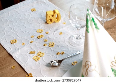 Stylish table setting prepared for a 50th birthday celebration. A golden number 50 centerpiece is placed on a white textured table runner, surrounded by matching gold confetti. Wine glasses, cutlery, and a decorative napkin complete the elegant dining arrangement. The image represents a milestone birthday, anniversary, or festive celebration in a refined and welcoming atmosphere. Suitable for themes such as birthdays, special occasions, dining, hospitality, and lifestyle events.