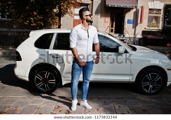 Stylish tall arabian man model in white shirt, jeans\
and sunglasses posed at street of city. Beard rich attractive arab\
guy against white suv\
car.