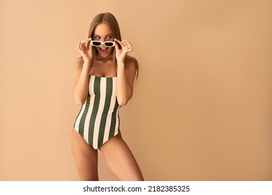 Stylish tanned woman in striped swimsuit and white sunglasses with surprise emotion on beige background.