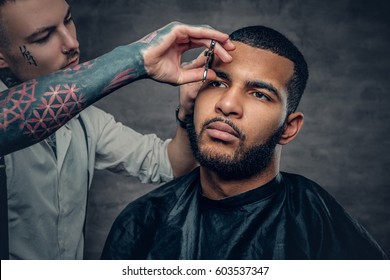 Stylish Tattooed Barber Makes Haircut To A Black Bearded Male.