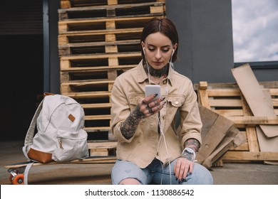 stylish tattooed woman in earphones listening music with smartphone near skateboard and backpack - Powered by Shutterstock