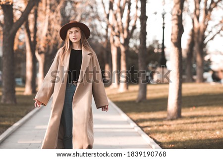 Stylish teen girl 13-14 year old wearing trendy beige coat and hat walking in autumn park. Fall season. Teenagerhood. 
