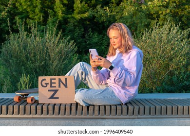 A Stylish Teenage Girl In A Lilac Hoodie Jeans With Pink Hair Sits On A Bench With A Phone In Hand Next To A Longboard And A Sign With The Inscription Generation Z. GEN Z STYLE  TRENDS.