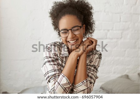 Stylish teenage girl wearing hipster eyeglasses and plaid shirt laughing, enjoying free time indoors, sitting on sofa against white brick wall background. Happiness, fun, joy, leisure and relaxation