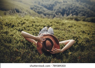 stylish traveler woman in hat lying on grass and relaxing in mountains. hipster girl on top of mountain, resting, hat on her face. space for text. atmospheric moment. wanderlust and travel concept.