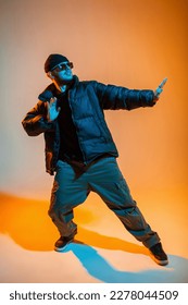 Stylish trendy handsome professional hip hop dancer man with sunglasses and cap in fashion black clothes with winter down jacket and sneakers dances in creative studio with orange and cold neon light - Shutterstock ID 2278044509