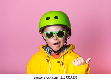 Stylish Trendy Kid Look. Eight Years Boy In Green Safety Helmet And Sunglasses And Yellow Bright Coat Isolated On Pink Background. Cool Portrait Of School Child