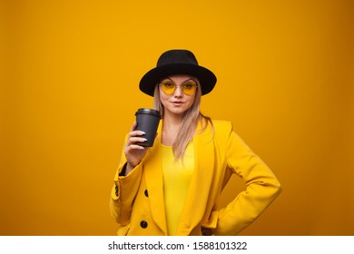 Stylish trendy young woman in bright clothes on a yellow background. Cool blonde girl in yellow jacket and black hat with a coffee to take away