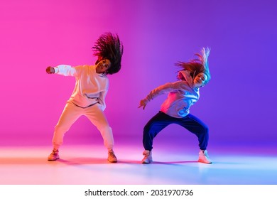 Stylish two beautiful young girls dancing hip-hop on gradient pink purple neon background. Sport achievement, spirit of expression. Concept of dance, youth, hobby, dynamics, movement, action, ad