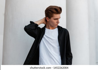 Stylish Urban Young Man In A Fashionable Black Shirt In A White T-shirt With A Stylish Hairstyle Stands Near A White Vintage Building With Columns In The City. Handsome Guy Fashion Model Outdoors.