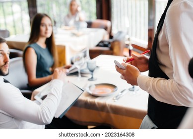 A Stylish Waiter Serves A Young Couple Who Came On A Date In A Gourmet Restaurant. Customer Service In The Catering.