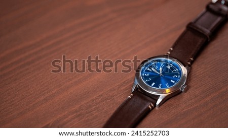 
A stylish watch with a brown leather band and a striking blue face rests gracefully on a wooden table, embodying timeless elegance and modern sophistication in this chic and fashionable timepiece