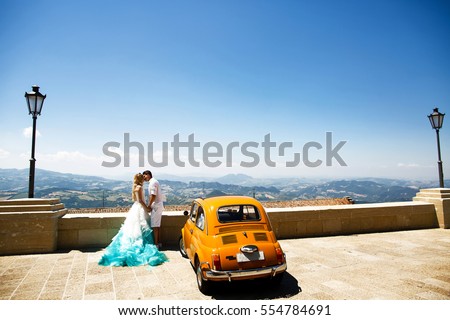 Stylish wedding couple kisses before yellow retro Fiat 500 with great view on mountains behind them