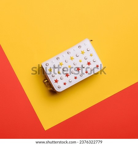Stylish white leather women's handbag covered with red, yellow, and white rivets in the shape of stars and rhinestones on a bright geometric background with copy space for text. Fashion blog content.