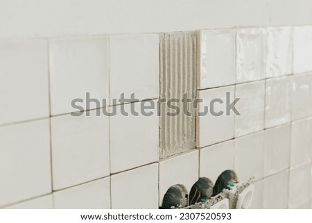 Stylish white tiles and electricity socket close up on wall. Modern square tile installation on adhesive and electric box. Kitchen or bathroom renovation, home improvement