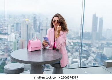 Stylish Woman In Beautiful Fashion Pink Blazer With Cute Bag Holding Mobile Phone On Rooftop Terrace With Amazing Bangkok View High Floor Industrial City