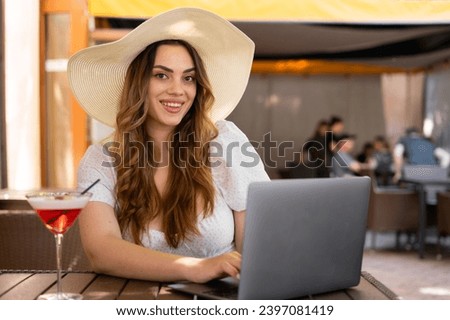 Stylish woman in big straw hat working remotely from a cafe, drinking cocktail and using laptop, smiling on camera. Freelance concept