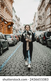 Mujer elegante en abrigo negro y bufanda caminando en calle empedrada - moda urbana, concepto de estilo de vida de la ciudad - peatón en paisaje urbano con coches aparcados, escena de invierno, exploración urbana y moda 