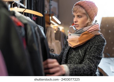 A stylish woman browses through winter coats in a boutique, reflecting on her fashion choices.