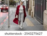 Stylish Woman in Burgundy Coat Walks City Street. Wearing Fashionable clothing White Turtleneck Sweater, Pearl Necklace and Grey Jeans, Carrying White Bag. Confident and Chic Mature Fashion Model