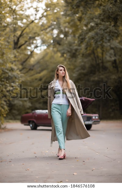 stylish woman in cape looking\
away while walking on road near broken retro car on blurred\
background