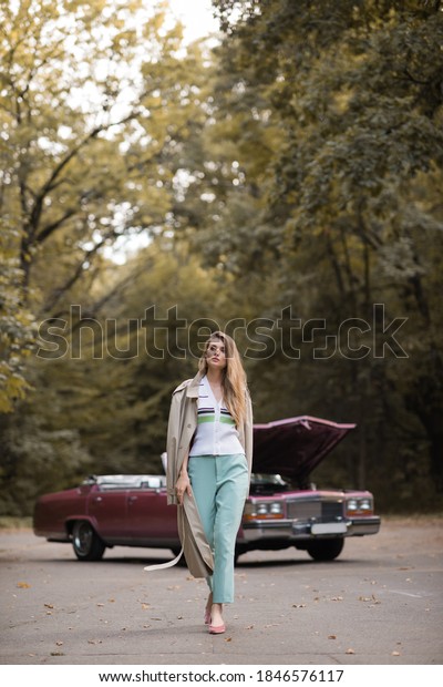 stylish woman in cape walking on road near\
broken vintage car on blurred\
background