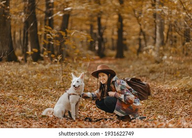 Stylish woman caressing adorable white dog in sunny autumn woods. Cute swiss shepherd puppy. Hipster female with backpack walking with her dog in autumn forest. Space for text