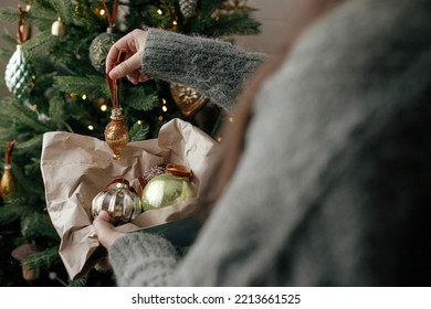 Stylish woman in cozy sweater decorating christmas tree with stylish bauble in atmospheric festive room. Merry Christmas! Winter holidays preparation. Decorating xmas tree with vintage toy