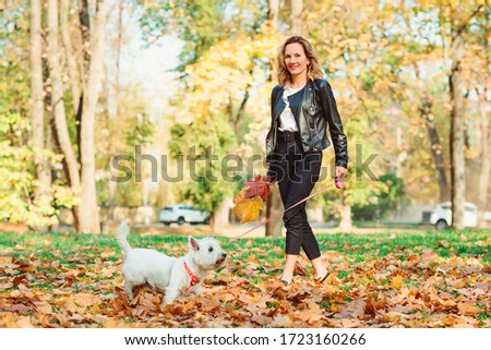 Stylish woman with dog on a walk in autumn park. Lifestyle, fashion and autumn mood concept. Friendship, animal and pet. Happy woman running with her little west highland white terrier in a park.