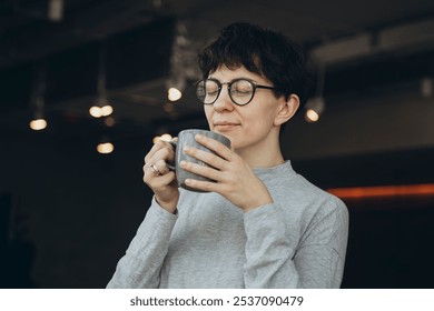 Stylish woman drinking coffee in the sunny morning in the office, morning routine. A woman with a short haircut and piercing smiles and drinks coffee. Successful woman on a coffee break. - Powered by Shutterstock