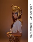 Stylish woman in Egyptian attire and headdress looking at camera while posing isolated on brown