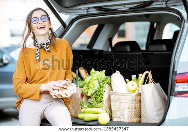 Stylish\
woman enjoying food while sitting on the car trunk full of healthy\
products on the supermarket parking\
outdoors