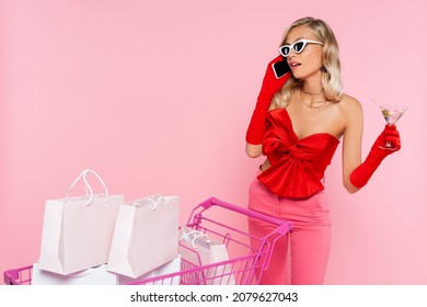 Stylish Woman With Glass Of Cocktail Talking On Mobile Phone Near Cart With Shopping Bags Isolated On Pink