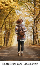 Stylish woman hipster in hat with backpack walking on road in sunny autumn woods. Young female traveler hiking in fall forest, beautiful moment. Travel and wanderlust concept. Back view